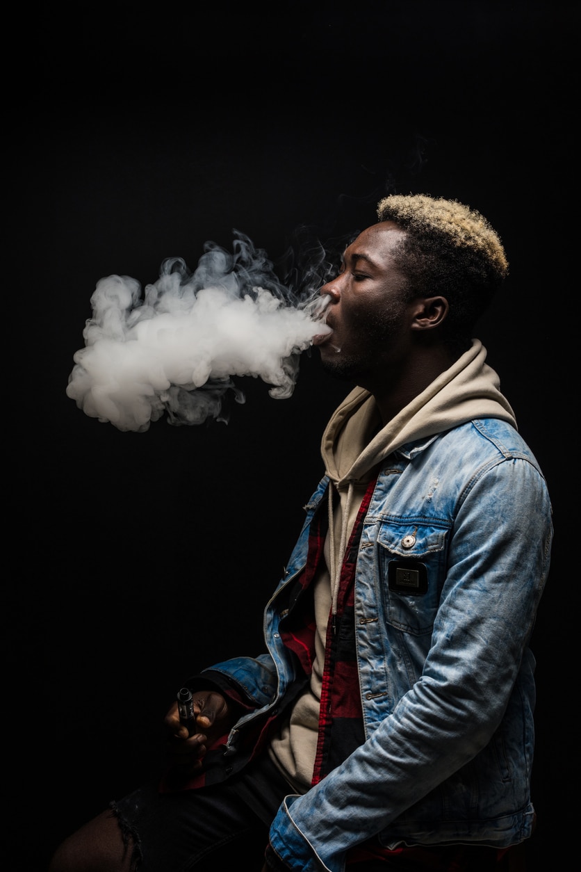 frican Man smoking or vaping e-cig or electronic cigarette holding a mod with a lot of clouds isolated on black background