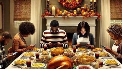 African American family with bowed heads at Thanksgiving dinner looking at devices - via EURai