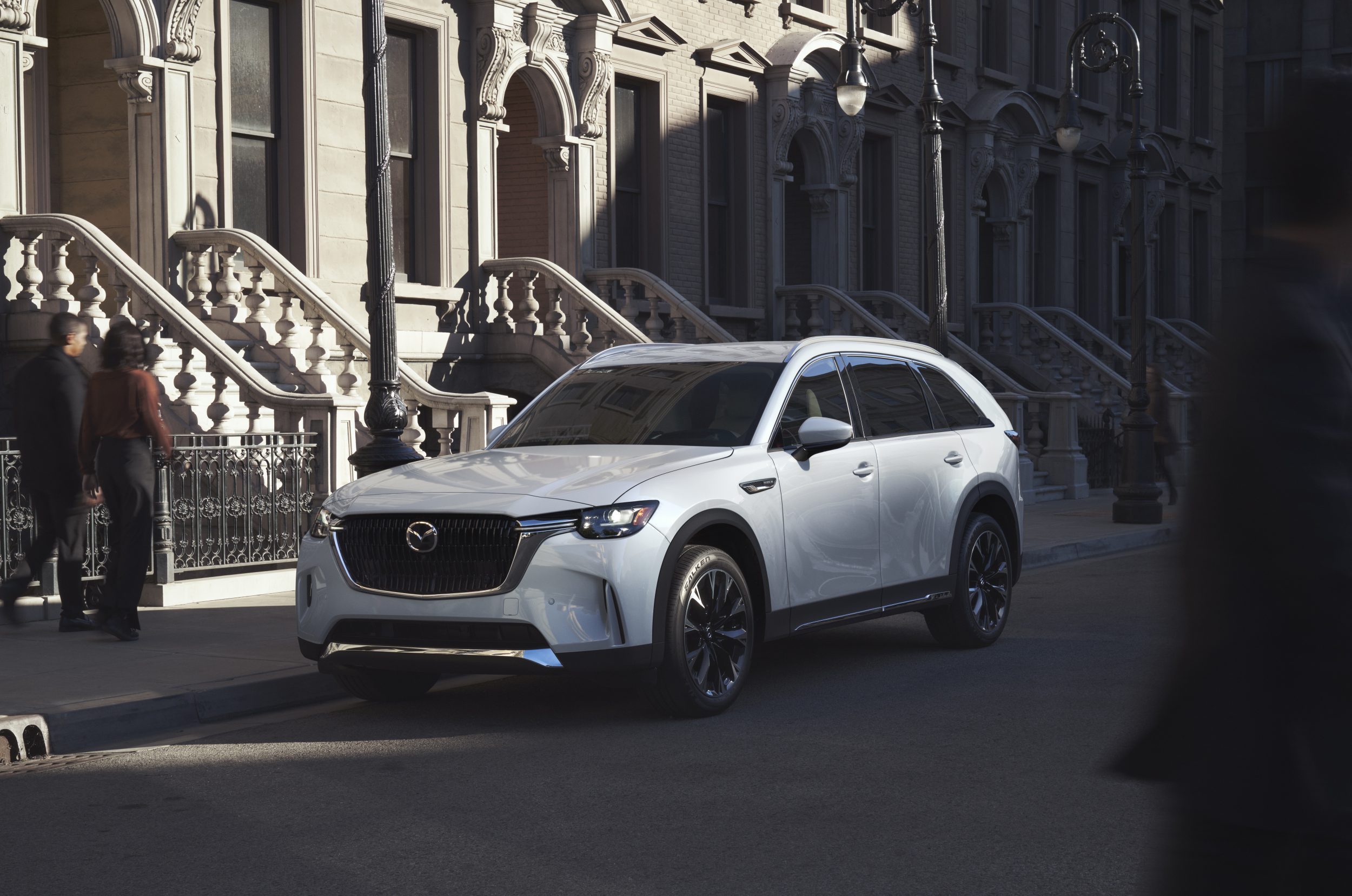 2025 Mazda CX-90 offers style, comfort & powerful trim options // white mazda suv pictured, parked along a curb beside stone architecture in the city