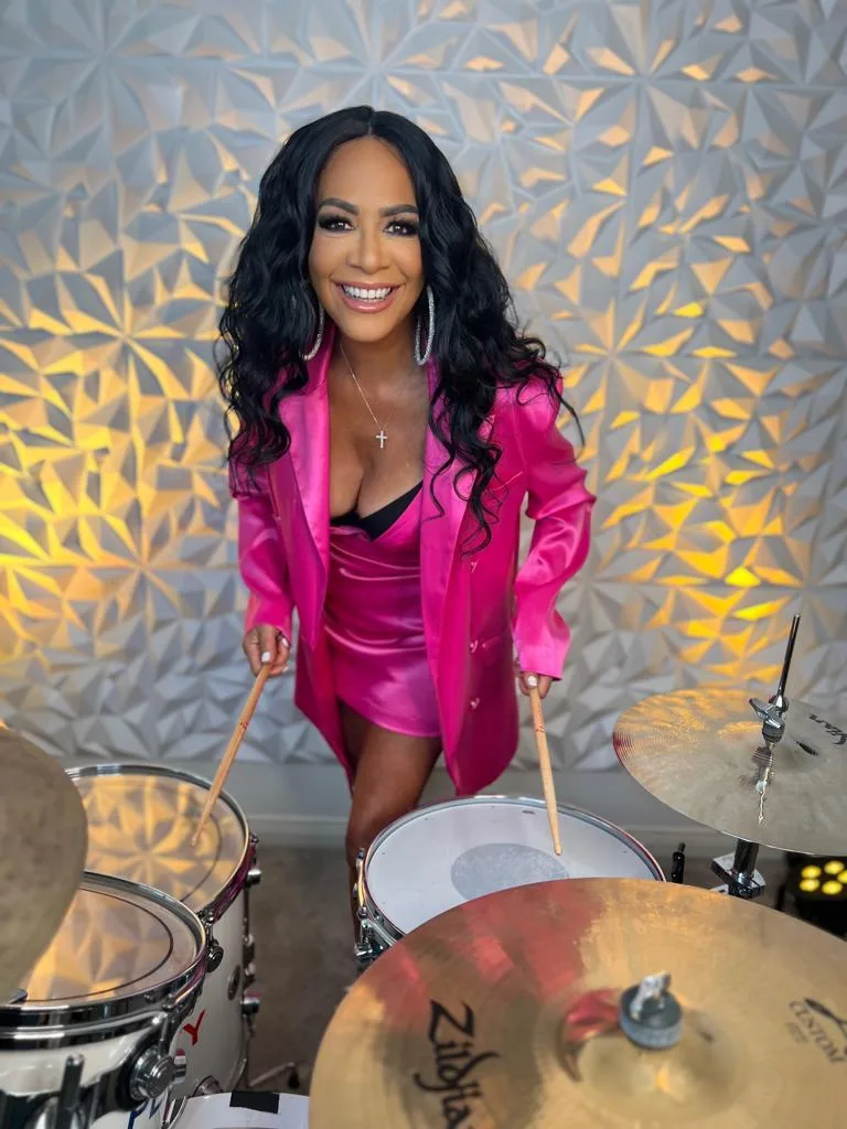 Sheila E. smiling in 2025 while standing behind a drum kit, wearing a hot pink blazer and satin dress.