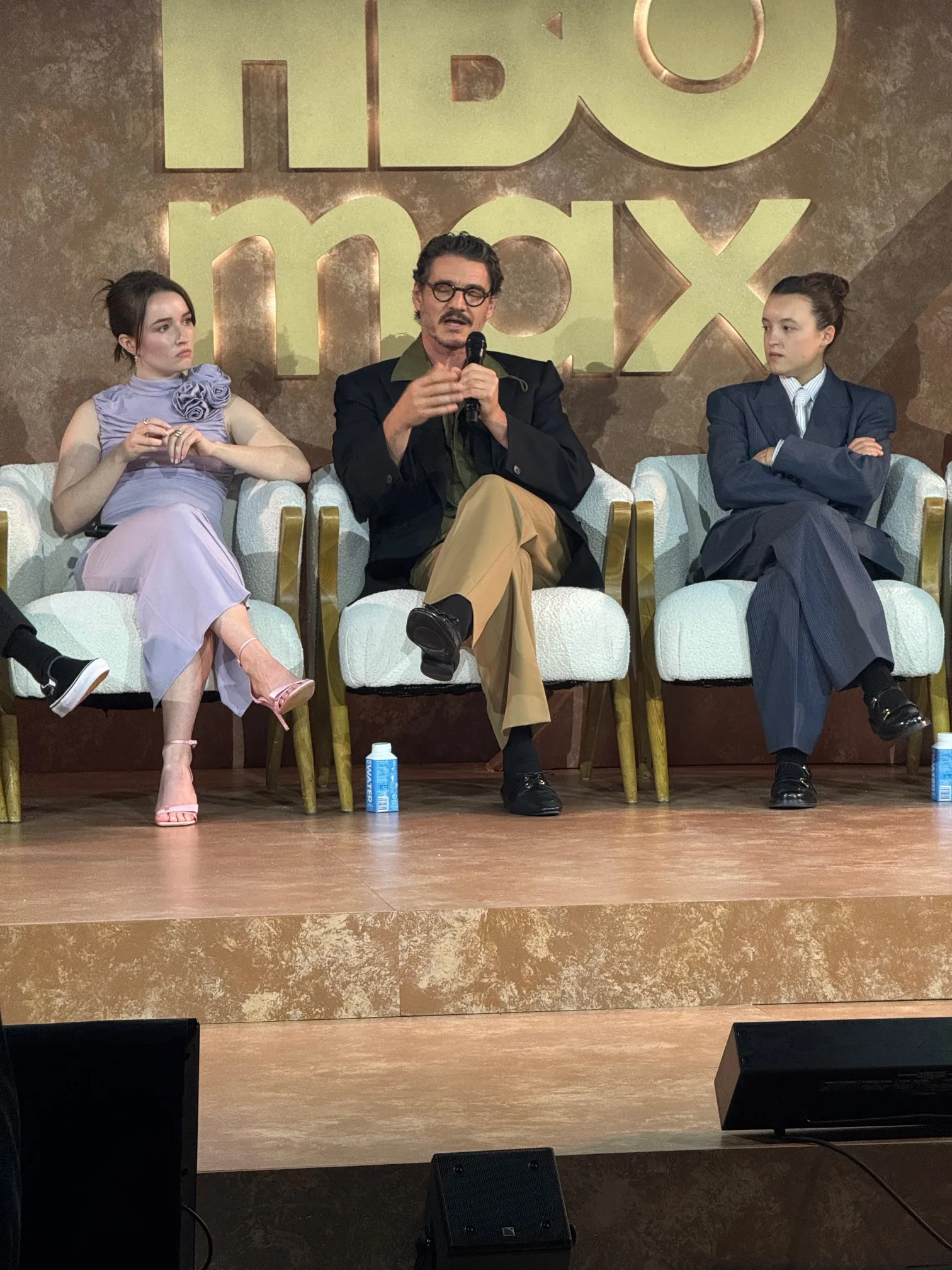 Pedro Pascal speaks into a microphone while Kaitlyn Dever and Bella Ramsey listen during an HBO Max Emmy event panel.