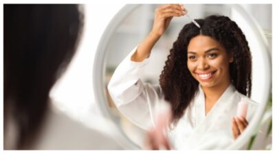 Smiling Black Woman Applying Serum For Hair Repair While Standing Near Mirror stock photo