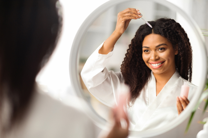 Smiling Black Woman Applying Serum For Hair Repair While Standing Near Mirror stock photo
