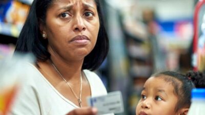 A worried mother at a grocery checkout holding her EBT card with minimal groceries and a small child beside her - via GrokAI