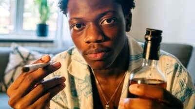 A young African American adult holding a cannabis joint and a closed bottle of alcohol, seated in a modern apartment, symbolizing a personal choice between weed and booze - via GrokAI