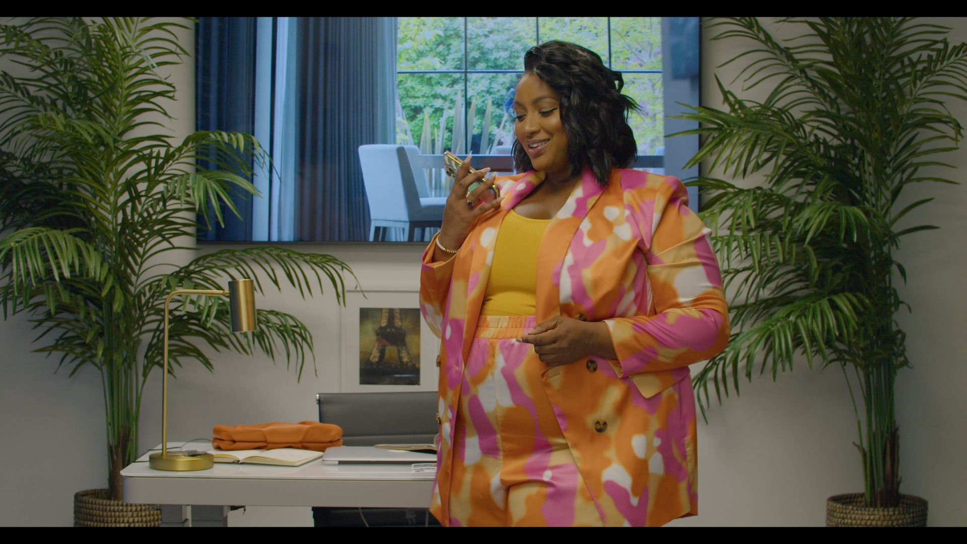 Nzinga Imani smiling while looking at her phone in a brightly colored suit inside a modern office setting.