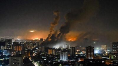 A nighttime scene in Caracas - smoke rising above buildings, with city lights flickering and distant flashes from explosions via GrokAI