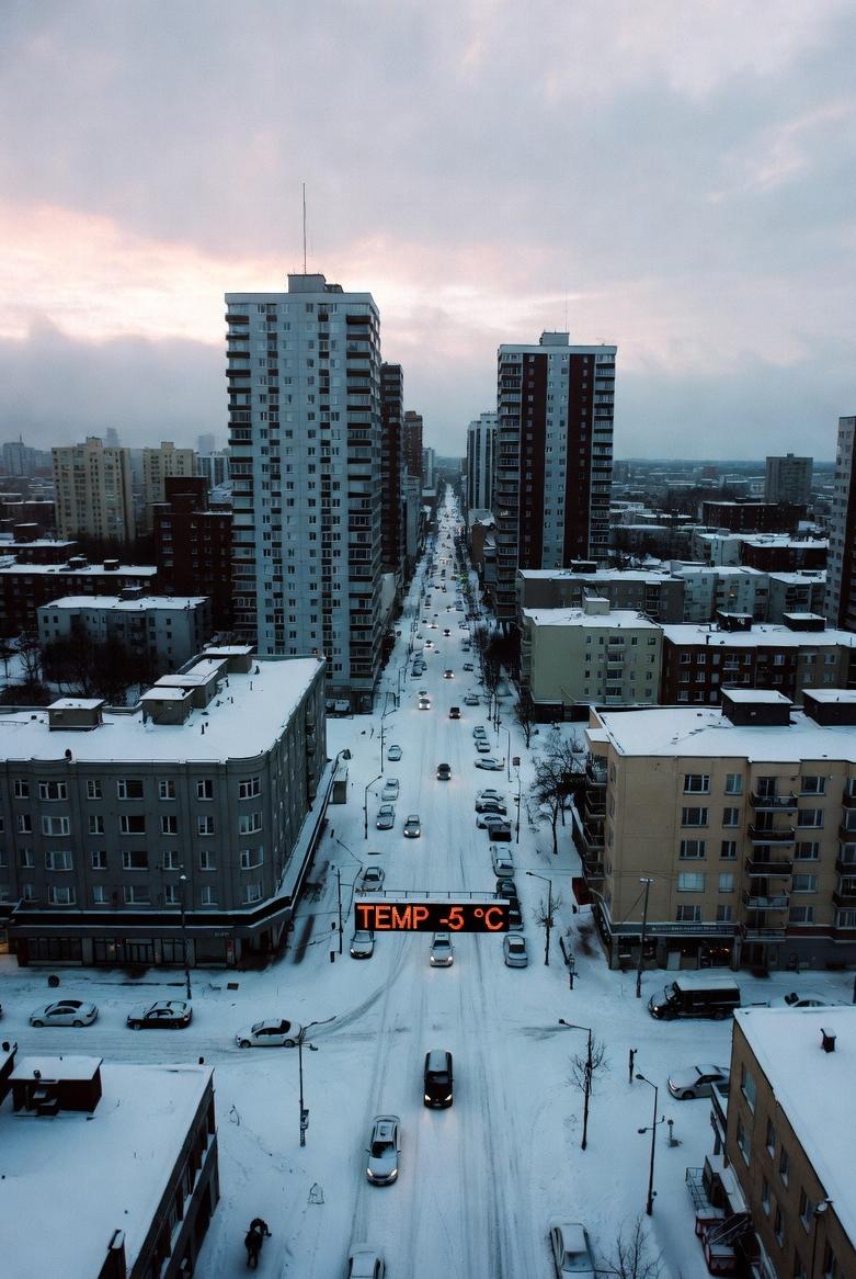 A snowy city-innercity at dawn, with thick snow covering rooftops and cars, and temperatures showing below zero - via eurAI