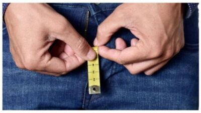 closeup of a young man holding a short piece of measuring tape that is popping up from the fly of his jeans