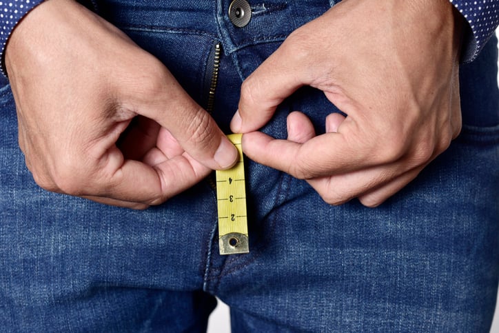 closeup of a young man holding a short piece of measuring tape that is popping up from the fly of his jeans