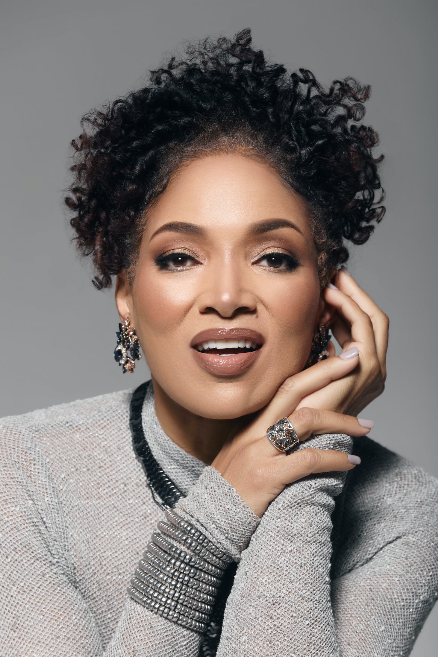 Lisa Arrindell poses in a studio portrait wearing a gray top with curly hair and statement jewelry