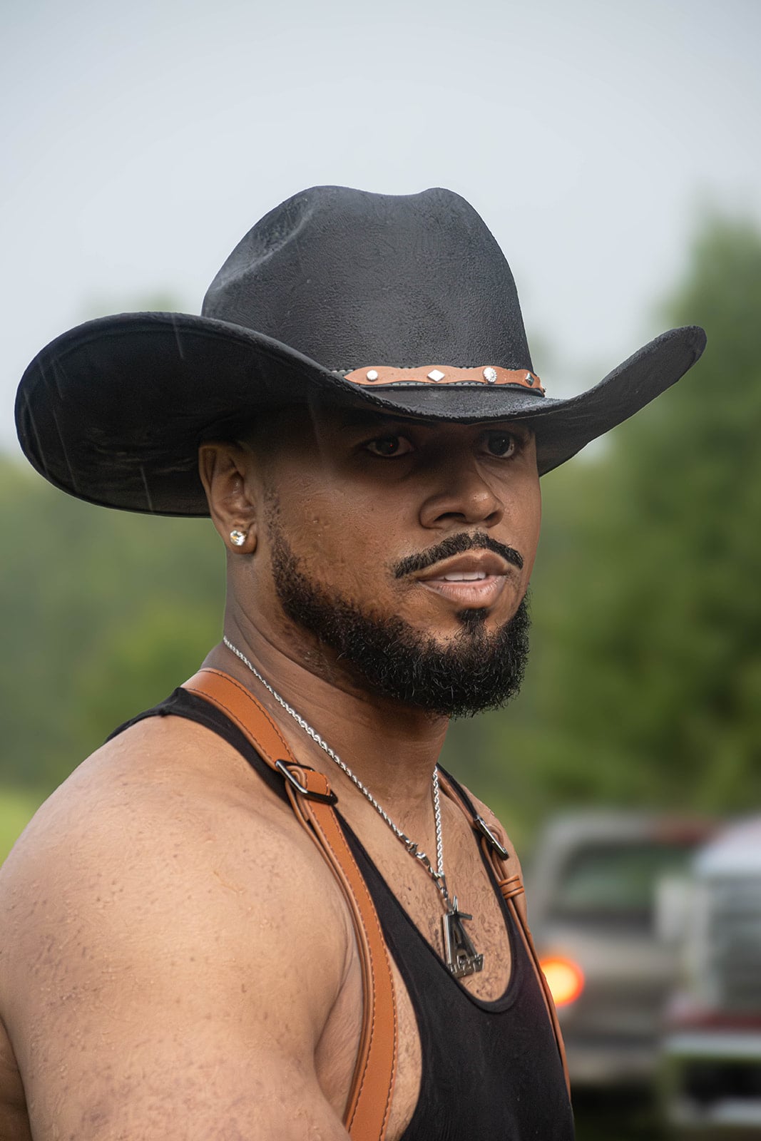 Julian Horton wearing a black cowboy hat, black tank top, and leather harness in an outdoor setting