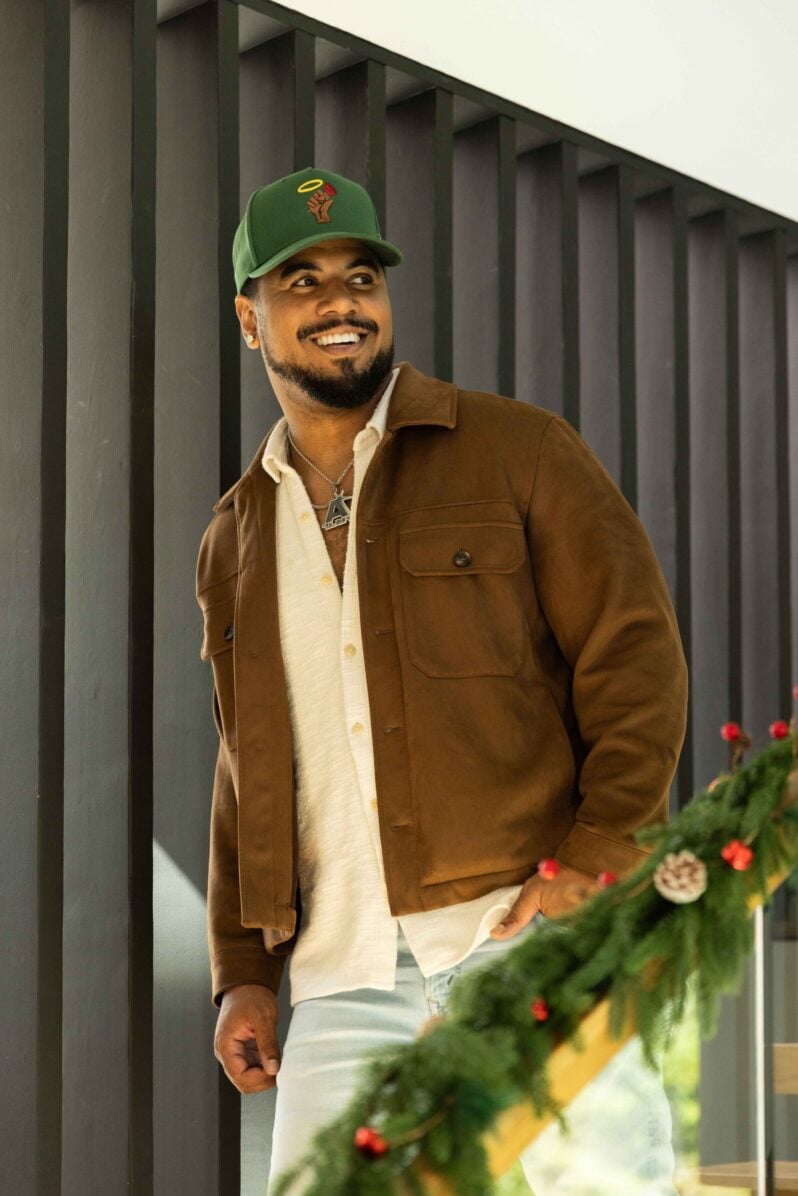 Julian Horton standing near a staircase wearing a green cap, brown jacket, white shirt, and light jeans