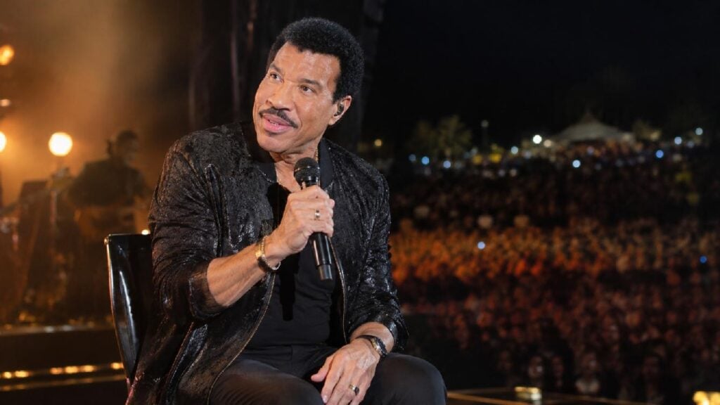 Lionel Richie seated onstage during a nighttime concert - via eurAI