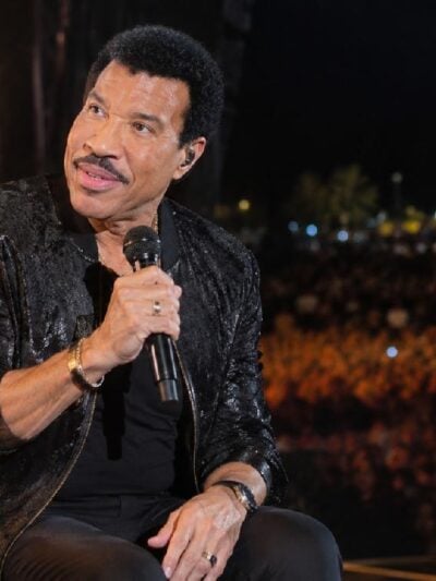 Lionel Richie seated onstage during a nighttime concert - via eurAI