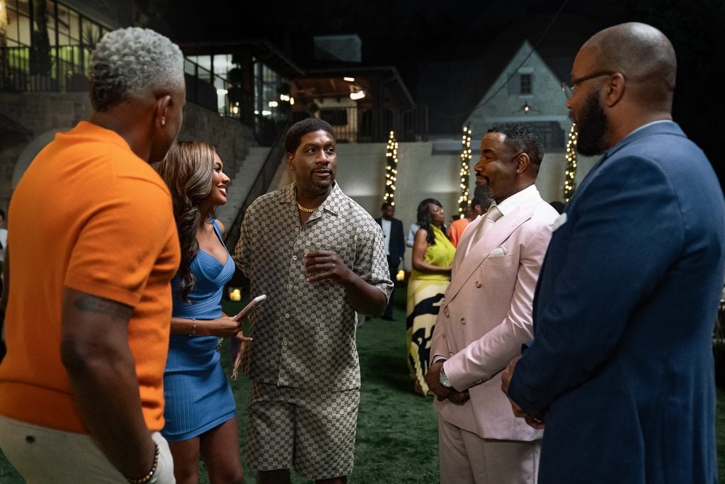 "Five adults dressed in stylish outfits stand talking and laughing at an outdoor evening party, with lights, greenery, and a large house visible in the background. The mood is friendly and festive."