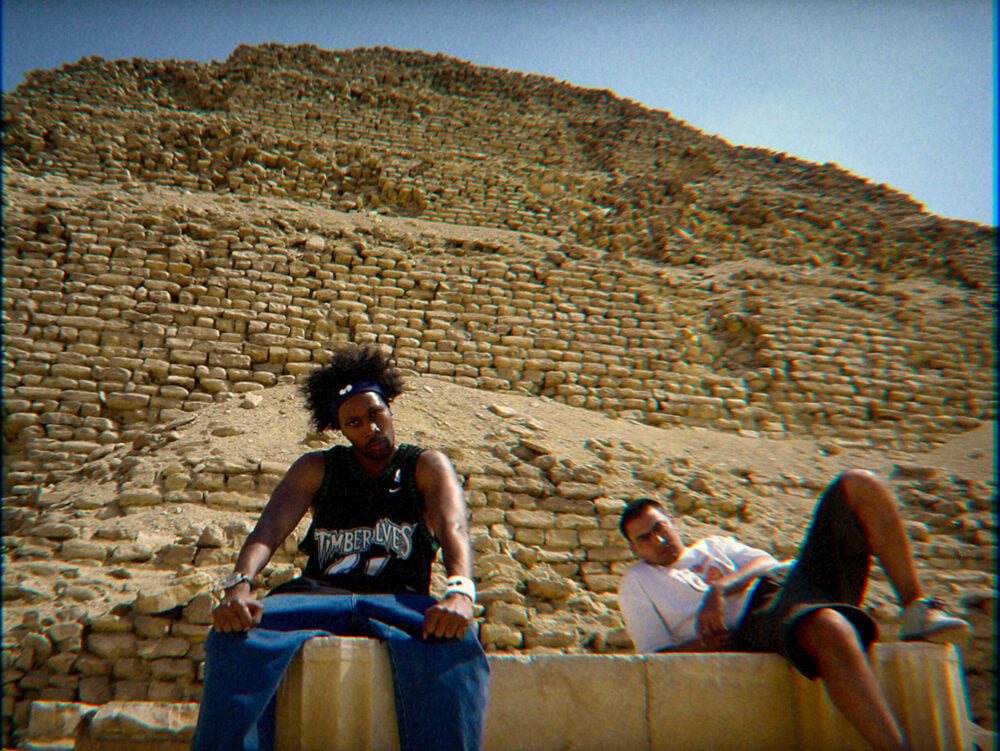 "Two men sitting relaxed in front of an ancient pyramid made of stone blocks under a clear blue sky, with one man wearing a basketball jersey and the other in casual clothing, both in a desert environment."